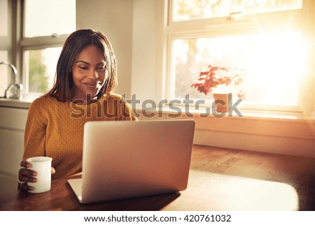 Adult female holding cup and laughing while looking at her laptop computer while sitting near bright sunny window in kitchen