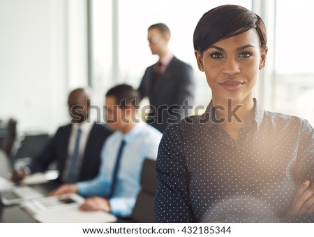 Attractive young grinning business owner in office with polka dot blouse, folded arms and confident expression in front of group of employees at conference table