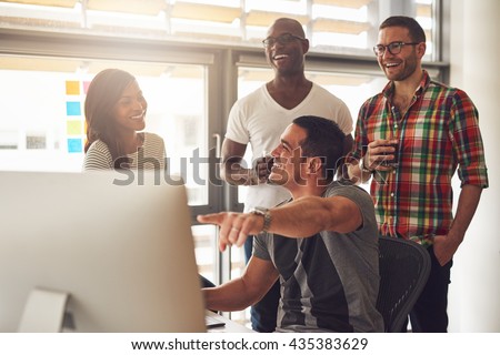 Handsome male adult pointing at something on his computer for a group of laughing male and female casually dressed friends holding drinks