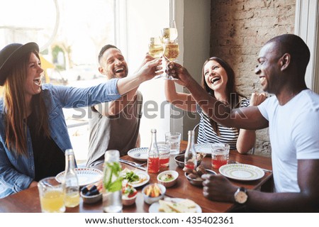 Group of diverse male and female laughing young adults putting their drinking glasses together at table in restaurant with large window
