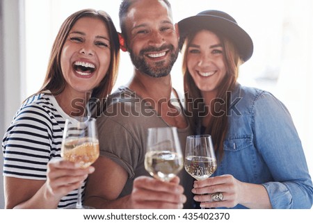 Three laughing friends in front of bright window light holding wine in glasses while celebrating something