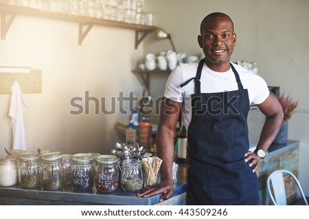 Handsome black entrepreneur stands by cafe counter lined with jars of tea while wearing dark colored apron