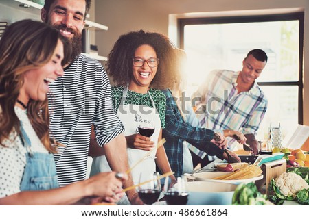 Group of diverse friends drinking and cooking in front of long food preparation table indoors