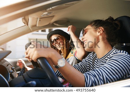 Two trendy attractive young woman singing along to the music as they drive along in the car through town viewed through the open side window
