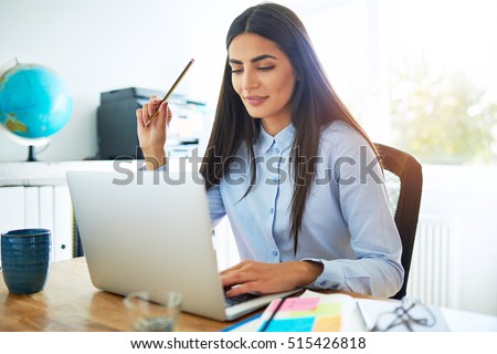 Single calm young Indian woman in blue blouse and long hair holding pencil in hand while seated at desk in front of laptop computer in bright room