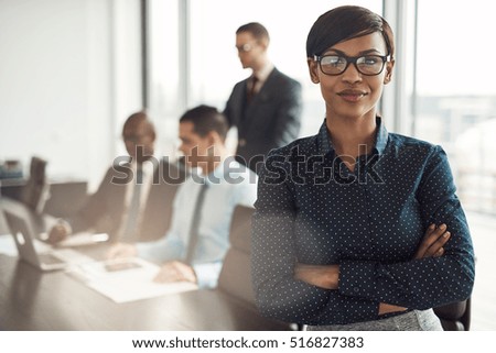 Confident young African businesswoman standing with folded arms smiling at the camera in a boardroom with male colleagues in the background