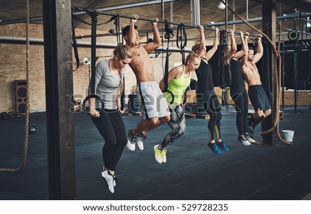 Group of six attractive young male and female adults doing pull ups on bar in cross fit training gym with brick walls and black mats