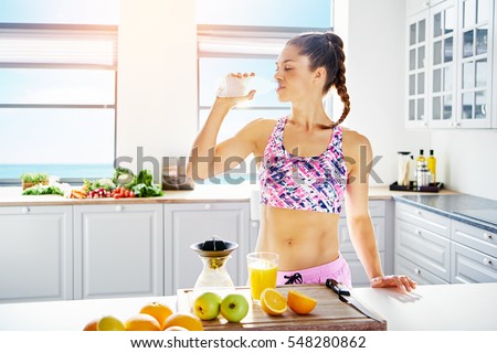 Young fit woman in sportswear drinking water while standing at kitchen.