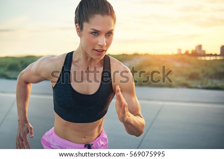 Fit determined young woman runner training at sunrise on a rural road sprinting towards the camera with a focused expression