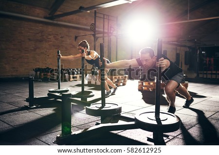 Muscular man and woman pushing hard bogie with weight disks. Horizontal indoors shot