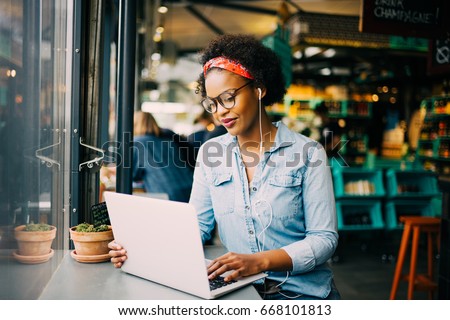 Focused young African woman sitting alone at a counter in a cafe working on a laptop and listening to music on earphones