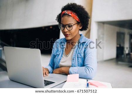 Smiling young African female entrepreneur hard at work on a laptop while sitting at a table in the lobby of a modern office building