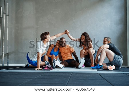 Two friends in sportswear high fiving each other while sitting on the floor of a gym talking with friends after a workout