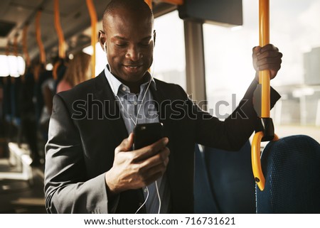 Smiling young African businessman wearing a suit standing on a bus during his morning commute listening to music on a smartphone and earphones