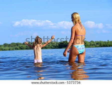 Beautiful mom in bikini looks at her daughter who wants to start swimming. Shot in June, near Dnieper river, Ukraine.