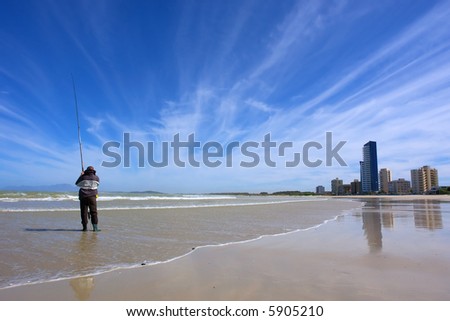Angler on beach with majestic sky and sky-scrapers. Shot in Strand, Western Cape, South Africa.