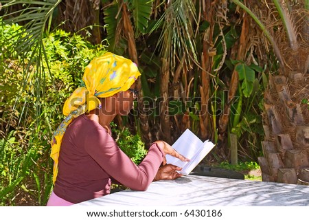 African woman in yellow turban reads book in shadow of a palm. Shot in the Botanical Garden, Stellenbosch, Western Cape, South Africa.