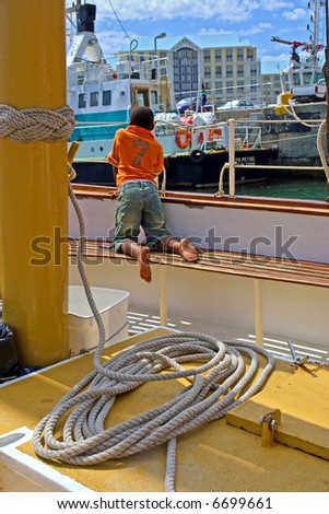 Black boy sits on yacht deck and looks at the pier. Shot during yacht cruise in Waterfront, Cape Town, South Africa.