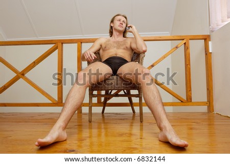 Relaxed man in trunks talks over phone sitting in chair. Shot in South Africa.