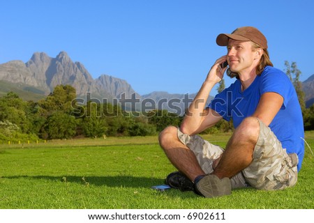 Smiling man sits on grass and talks over phone - and misty mountains are background. Shot in Stellenbosch, South Africa.