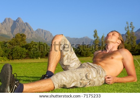 Young man sunbathing on grass in sunset light - and beautiful mountains are background. Shot in Stellenbosch Mountains, Western Cape, South Africa.