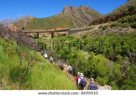 Long file group of hikers walks towards highway bridge across small river in awesome mountains. Shot in the Kromrivier - Du Toitskloof Nature Reserve, near Paarl, Western Cape, South Africa.