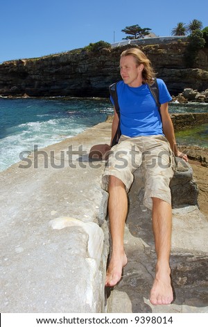 Happy young man sits on stone next to sea. Shot in Hermanus, Walker Bay, Western Cape, South Africa.