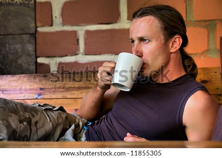 Young man drinks coffee lying next to fire - shot against red brick wall. Shot in Sodwana Bay, KwaZulu-Natal, South Africa and Southern Mozambique.