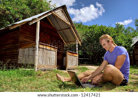 Smiling man with laptop on grass next to rural house. Shot in Sodwana, KwaZulu-Natal province, South Africa.