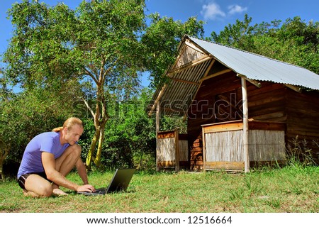 Busy man with laptop squats on grass next to rural house. Shot in Sodwana, KwaZulu-Natal province, South Africa.