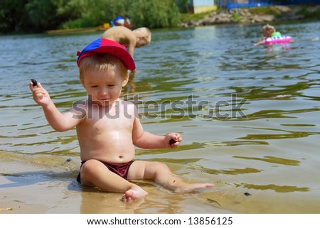 Smiling baby boy throws stones into water sitting on beach. Shot in July, Ukraine.
