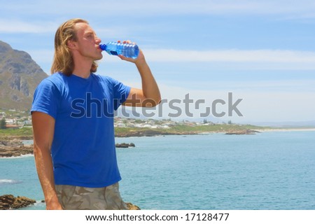 Young man drinks from water bottle standing on rock next to sea. Shot on Cliff Path near Hermanus, Walker Bay, Western Cape, South Africa.