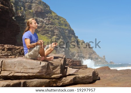 Young man meditates on rocks - sea-misty mountain background. Shot in the Cape of Good Hope and Cape Point Nature Reserve, Table Mountain National Park, near Cape Town, South Africa.