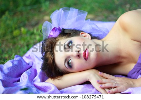 Cute young brunette female lying on grass field at the park on the piece of violet fabric