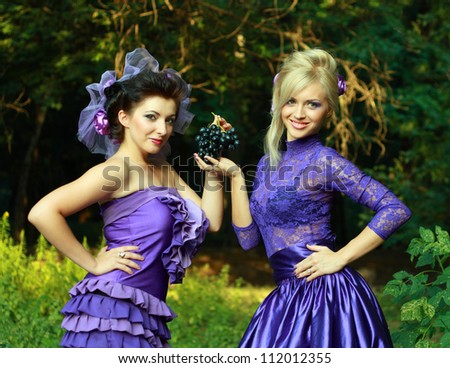 two fashionable beautiful women both in violet dresses holding bunch of grapes and smiling