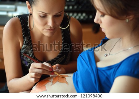 body art make up artist creating flower painting on the belly of pregnant young woman