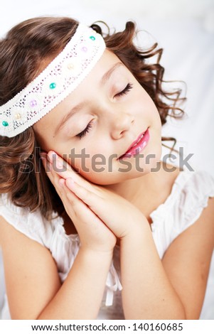 Cute smiling little girl with red hair showing it is time to sleep with her hands near cheek, isolated on white