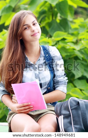 Beautiful school or college girl sitting on the bench with book and bag studying in a park