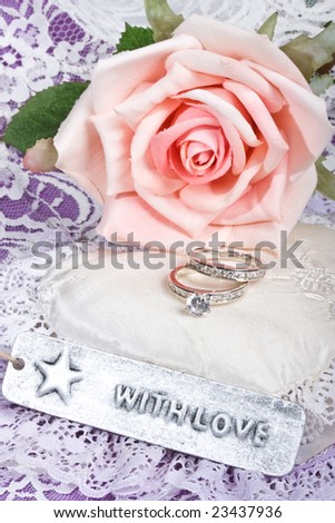 'with love' tag in front of pink rose and rings on satin and lace heart pillow