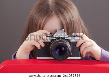 Young caucasian child with a retro cam in her Hand take a picture behind a red chair.
Finger on the shutter.