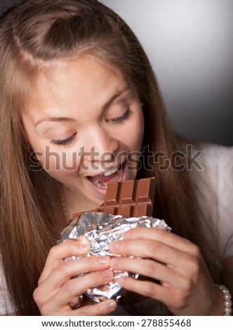 Close up Pretty Girl Eating Chocolate Bar in Foil While Looking at it, with Mouth Wide Open. Captured in Studio with Gray Gradient Background.