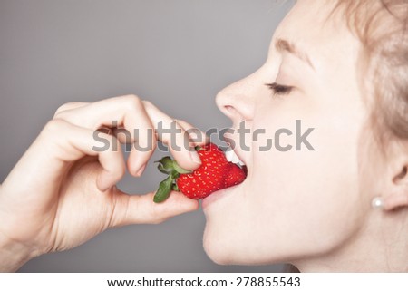 Close up Pretty Girl Biting a Fresh Red Strawberry Fruit in a Sexy Way with Eyes Closed Against Gray Background, Captured in Side View.