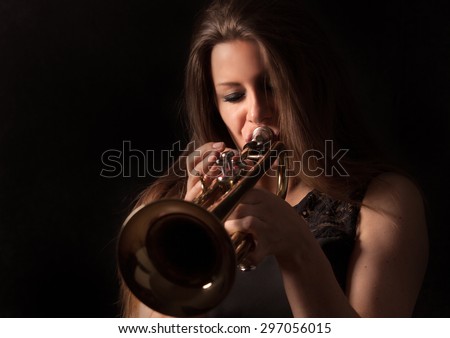 Women with brown hair is playing a trumpet in front of black background