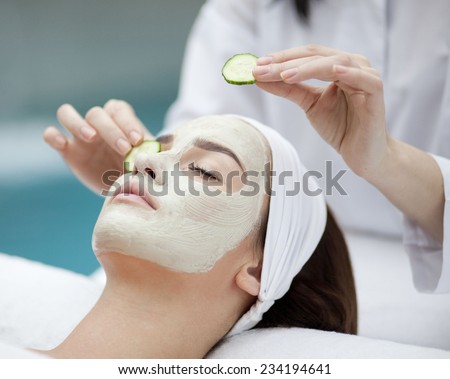 Beautiful young woman receiving facial mask of cucumber in beauty salon, hands of cosmetologist
