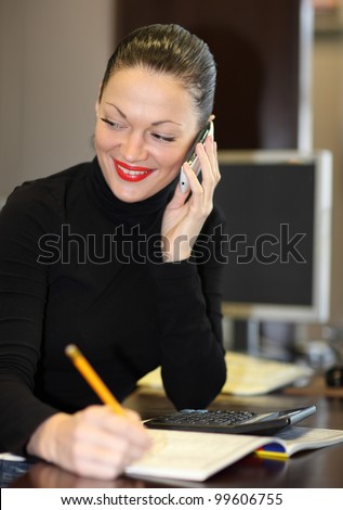 Happy office girl at desk working on desktop computer, using phone, smiling.
