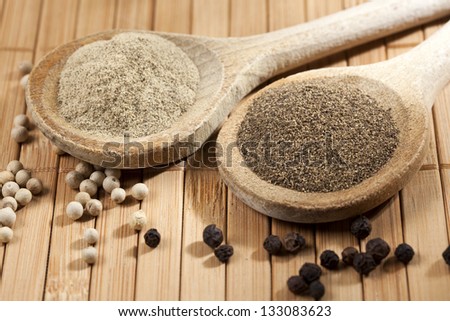 white and black pepper as peppercorns and heaps of powder on wooden spoons