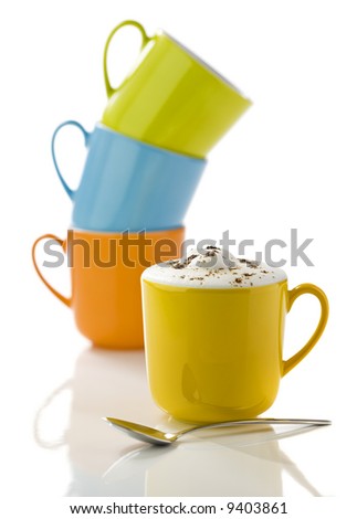 yellow mug filled with cappuccino, spoon in front, green, orange and blue mugs stacked in background, on white reflective surface