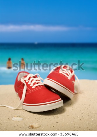 Red sneakers on beautiful beach. Two persons walking into turquoise water. Shallow depth of field, focus on the sneakers. Digital composite.