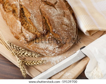 loaf of handcrafted whole wheat bread with wheat stalks, knife, towel, flour, view from above