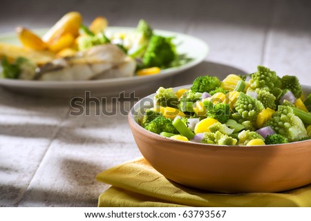 various steamed vegetables in terracotta bowl on table, plate with grilled fish out ot focus in background